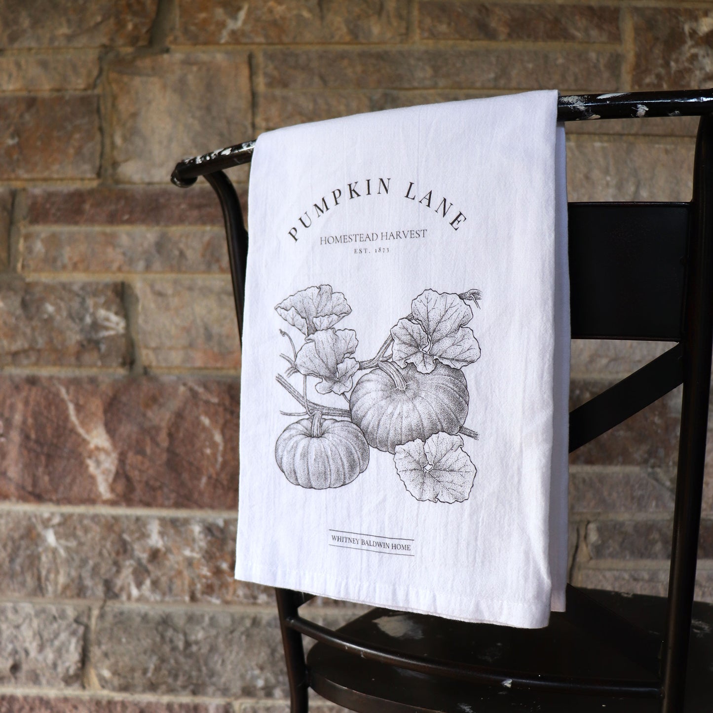 Homestead Harvest Pumpkin Lane Tea Towel. The Pumpkin lane fall kitchen towel is hung over the back of a chair