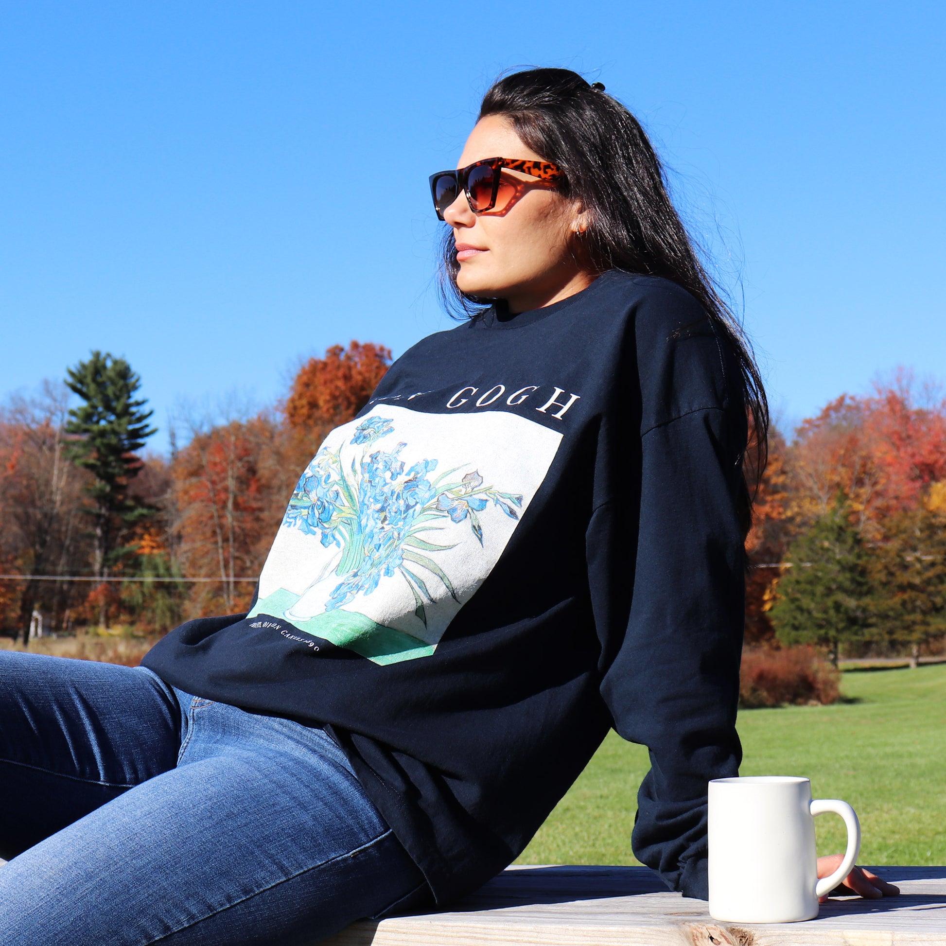 woman wearing navy long sleeve vincent van gogh tshirt sitting on a picnic table with a cup of coffee looking off into the sunset