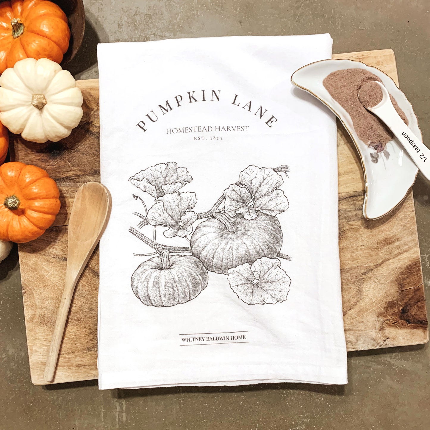Homestead Harvest Pumpkin Lane Tea Towel. This tea towel is displayed with mini pumpkins on a cutting board with spatula and cocoa mix. The pumpkin illustration is of a small pumpkin patch 