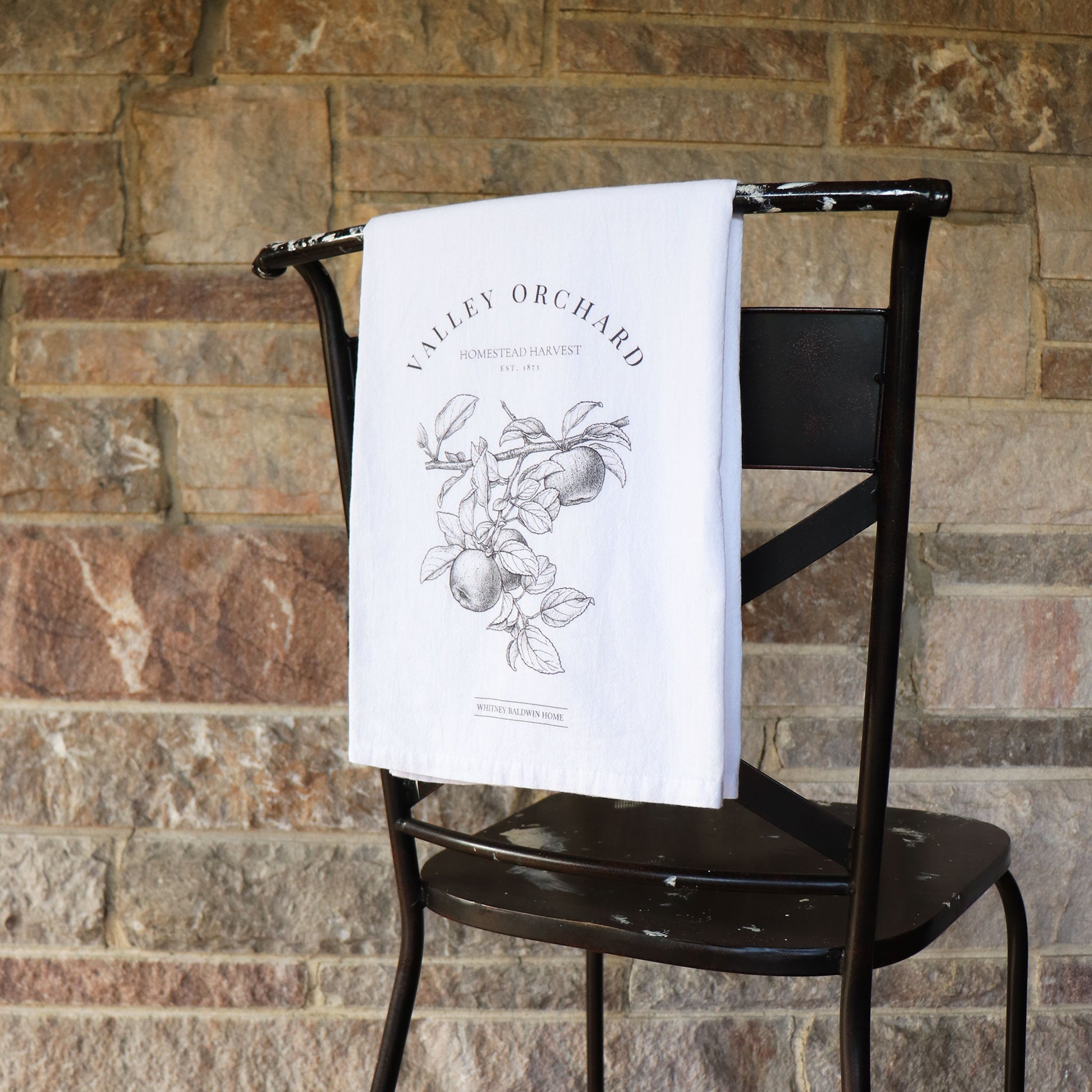 homestead harvest valley orchard apple orchard tea towel. the apple orchard tea towel is hung over the back of a char. The illustration displays a branch with two apples on it.