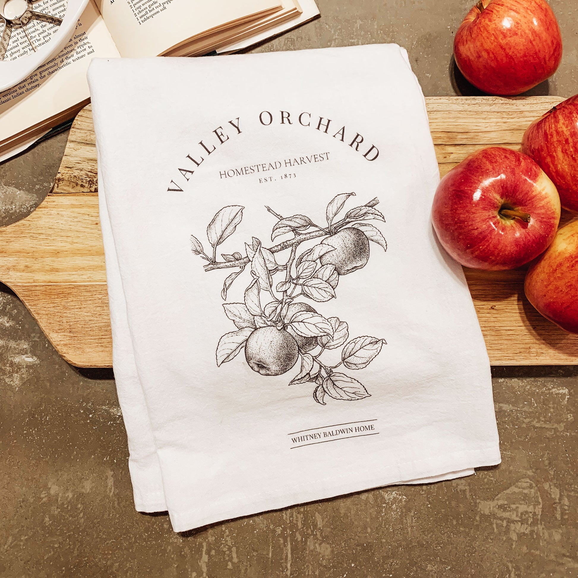 homestead harvest valley orchard apple orchard tea towel. Tea towel displayed with cutting board, apples, cook book and apple slicer